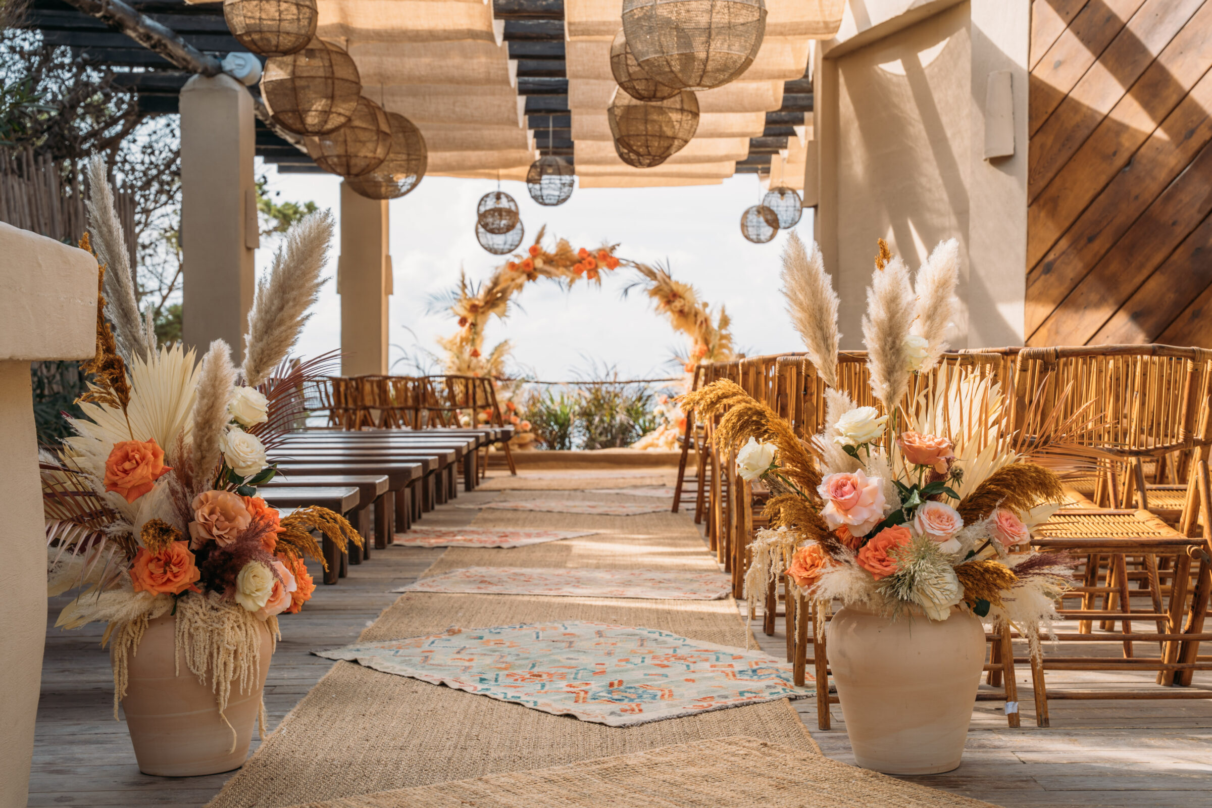 Ceremonia de boda al aire libre con arco floral en tonos tierra, beige, coral y rosa, rodeado de vegetación mediterránea.
